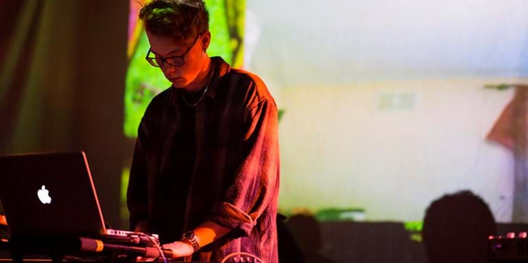 Male student performing a DJ set on an Apple Macbook, wearing a long sleeved shirt, in shadow against a projection background.