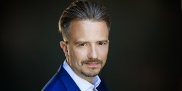 Andrew Griffiths - headshot on black. Male, wearing a blue suit, head and shoulders.